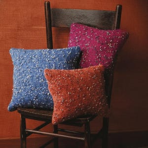 May include: Four textured cushions in shades of blue, red-orange, and magenta sit on a dark wooden chair. The cushions have a speckled pattern. A bouquet of purple flowers lies on the floor. The image includes the text "KNITTING PDF PATTERN".
