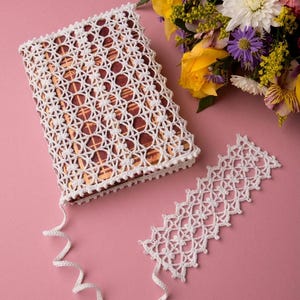 Könnte beinhalten: Ein rechteckiges Buch in einem weißen Häkelumschlag mit wiederkehrendem geometrischen Muster, dazu ein passendes weißes Lesezeichen aus Häkelarbeit. Beide Teile haben eine weiße Kordel mit weißer Quaste und einem kleinen goldenen Metallakzent.
