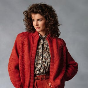 May include: A vibrant red knit cardigan featuring a textured diamond pattern on the sleeves and front panels, paired with a button-down shirt with a brown and cream paisley print and a matching red skirt with a thin brown leather belt.