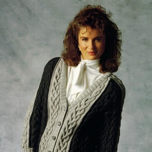 May include: A V-neck button-down cardigan featuring a color-blocked design with black and light gray panels. The garment is crafted with a prominent cable knit texture, ribbed cuffs, and a ribbed hem, worn over a white blouse with a tied neck detail.