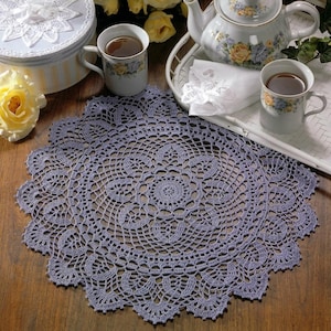 May include: A round, lavender-coloured crochet doily featuring an intricate, openwork floral pattern with a scalloped edge. The doily is displayed on a wooden surface alongside a white tea set, yellow roses, and a round, light blue decorative box.