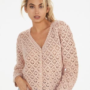 Può includere: Un cardigan a maniche lunghe rosa chiaro caratterizzato da un intricato motivo a uncinetto traforato con fiori ripetuti. Il capo presenta uno scollo a V, un bordo smerlato lungo l'apertura frontale e si chiude con una fila di piccoli bottoni rotondi chiari.