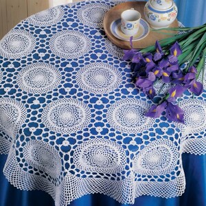 May include: A white round crochet tablecloth featuring a repeating circular motif pattern with an intricate openwork design. The lace fabric is draped over a solid blue surface, creating a textured contrast against the smooth fabric underneath.