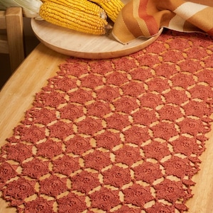 May include: A rust-colored crochet table runner featuring a repeating star-shaped motif pattern. The runner is laid out on a light-toned wooden surface, with a wooden bowl containing three ears of corn and a striped fabric napkin nearby.
