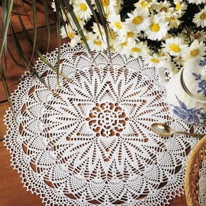 Puede incluir: Un tapete de ganchillo blanco redondo que presenta un patrón de estrella intrincado y simétrico con delicadas puntadas caladas. El tejido circular muestra un motivo floral central rodeado de segmentos geométricos radiantes.