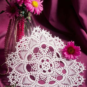 Op de afbeelding: Een rond wit gehaakt kleedje met een ingewikkeld opengewerkt patroon en een geschulpte rand, weergegeven naast een heldere glazen vaas gevuld met levendige roze madeliefachtige bloemen en een streng witte parels op een dieppaarse stof.