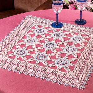 May include: A square crochet doily featuring a repeating floral pattern in light blue and cream white thread. The intricate lace design includes a scalloped border and openwork motifs, centered on a solid pink tablecloth with two stemmed glasses.
