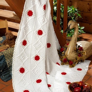 Puede incluir: Una manta de ganchillo blanca con un patrón repetitivo de diamantes con puntos de relieve y pequeños acentos florales rojos circulares. La manta tiene un borde festoneado y está drapeada verticalmente, mostrando su textura.