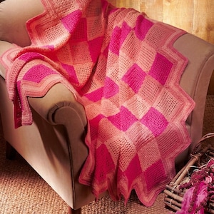 May include: A crochet blanket featuring a geometric pattern of alternating squares in vibrant shades of hot pink, light pink, and peach. The blanket has a distinct zigzag edge and a textured, woven appearance throughout the entire piece.