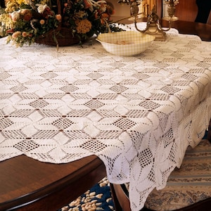 Puede incluir: Un mantel blanco, intrincadamente tejido a ganchillo, con un patrón geométrico, cubre una mesa de madera oscura. Una cesta de mimbre llena de flores y un cuenco a cuadros amarillos y blancos están sobre la mesa. El texto "PDF File PATTERN" es visible.