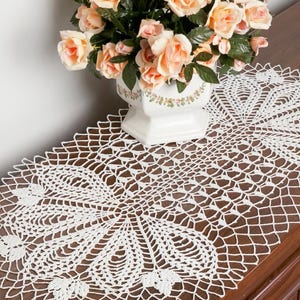 May include: A white oval crochet doily featuring an intricate openwork pattern with repeating floral-inspired motifs and a delicate scalloped edge, displayed underneath a white ceramic vase filled with peach-colored artificial roses.