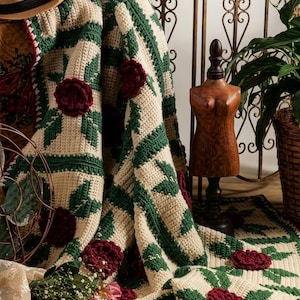 Può includere: Una coperta all'uncinetto color crema con un motivo ripetuto di foglie verde scuro e applicazioni di rose tridimensionali rosso intenso, rifinita con un bordo rosso scuro e drappeggiata su un manichino in legno.