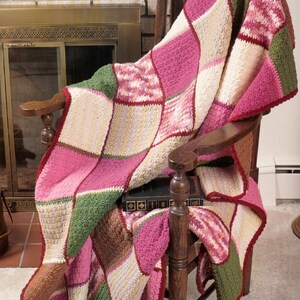 May include: A handmade patchwork crochet blanket draped over a wooden chair. The blanket features squares of pink, green, cream, and brown, with a red border. The blanket is a cozy home decor item.