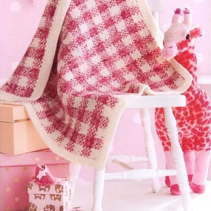 Może przedstawiać: Dzianinowy kocyk dla dziecka z czerwono-białym wzorem w kratę jest zarzucony na białym krześle. Pluszowa zabawka żyrafa z czerwono-białym wzorem w plamki stoi za krzesłem. Różowe pudełka na prezenty i buciki dla dziecka na pierwszym planie.