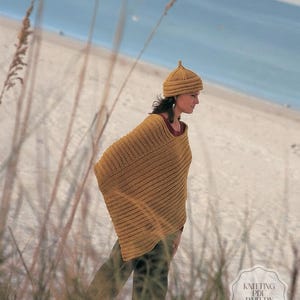 May include: A mustard yellow knitted poncho and matching hat. The poncho has a ribbed design and is worn over a dark red top. The hat has a pointed top. The background shows a beach scene with tall grass in the foreground.