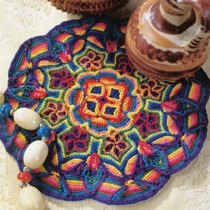 Op de afbeelding: Een levendige, regenboogkleurige gehaakte onderzetter met een bloemenpatroon. De onderzetter heeft een donkerblauwe rand en een centraal vierkant patroon. Een bruine keramische kan en een kralenketting zijn ook zichtbaar.