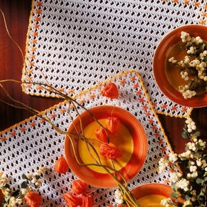 May include: Two rectangular white crocheted placemats with an openwork pattern, featuring a yellow crocheted border and orange cross-stitch accents. The placemats are arranged with orange and yellow ceramic plates and dried orange physalis.