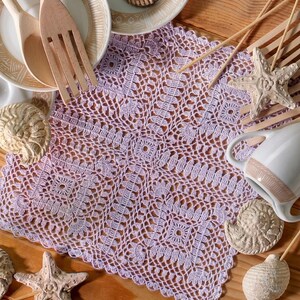 May include: A square lavender crochet doily featuring an intricate geometric and floral pattern. The doily is surrounded by wooden utensils, ceramic plates, a white pitcher, and several decorative seashells and starfish on a wooden surface.