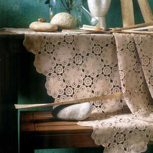 Puede incluir: Un mantel de ganchillo beige con un motivo floral circular repetitivo y un borde festoneado. El tejido está drapeado sobre una superficie de madera, con piedras lisas y un listón de madera encima. El texto PDF File Pattern es visible.