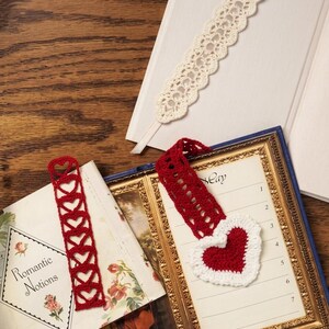 Puede incluir: Tres marcapáginas de ganchillo: una tira de encaje color crema con cinta, una tira roja con una fila vertical de corazones calados y una pieza roja en forma de corazón con borde blanco unida a una banda roja, sobre libros abiertos.