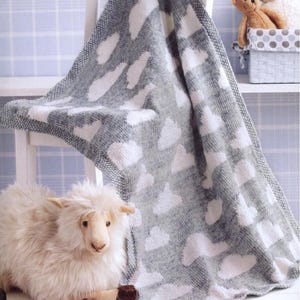 May include: A gray and white knitted baby blanket with a cloud pattern is draped over a white chair. A plush sheep toy and a basket with teddy bears are in the background. The blanket is approximately 40 inches by 50 inches.