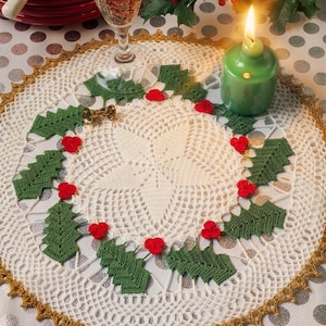 Op de afbeelding: Een rond, gebroken wit gehaakt kleedje met een centraal sterpatroon en een decoratieve goudkleurige rand. Het kleedje is versierd met verschillende kleine, gehaakte groene hulstblaadjes en groepjes van drie rode bessen in een cirkel.