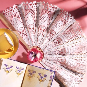May include: A white crochet hand fan with intricate lace patterns, featuring a pink pansy flower at the base. The fan is displayed alongside a yellow cup on a saucer and an open book with illustrations of purple and yellow pansy flowers.