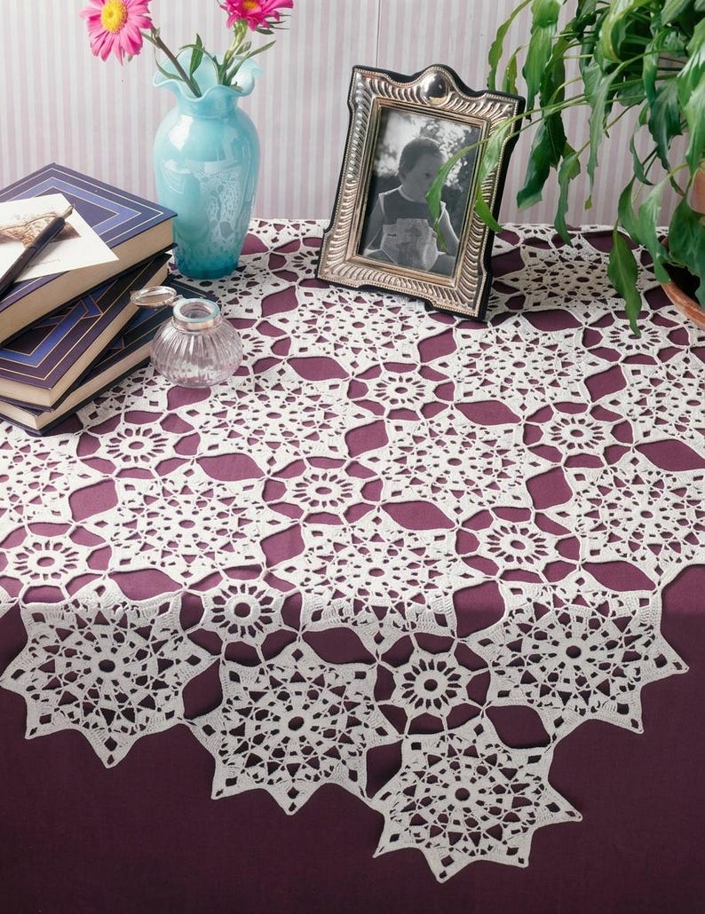 May include: A white crocheted tablecloth featuring a repeating star-shaped motif with intricate openwork patterns. The fabric is draped over a dark purple surface, showcasing a delicate, interconnected lace design with pointed edges and circular centers.