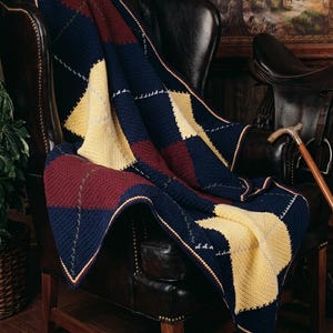 Op de afbeelding: Een gebreide plaid met een argyle-patroon in marineblauw, bordeauxrood en lichtgeel. De deken is gemaakt met een textuursteek en bevat dunne, lichtgekleurde diagonale lijnen die elkaar kruisen over de ruitvormen.