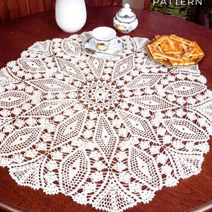 Puede incluir: Camino de mesa redondo de ganchillo color crema con un patrón floral detallado. El camino de mesa está sobre una mesa de madera oscura, con una taza y platillo, un azucarero pequeño y un plato de galletas. El texto "PDF File PATTERN" es visible.