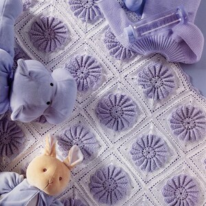 Könnte beinhalten: Eine weiße gehäkelte Decke mit lavendelfarbenen Blumenmotiven, begleitet von einem lila Plüsch-Nilpferd, einem Plüschhasen mit hellblauer Jacke, einer lavendelfarbenen Strickmütze und einer transparenten Babyflasche aus Kunststoff.