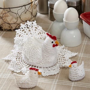 May include: A white crocheted hen with a lacy tail and two smaller white crocheted chicks. Each bird features a red comb, a red wattle, and an orange beak. The hen sits on a circular white doily, and the chicks are positioned in front of it.