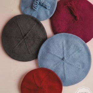 May include: A collection of knitted berets in various colors, including blue, burgundy, gray, and red. The berets are arranged in a circular pattern, showcasing their round shape and textured knit design. Some berets have decorative elements like a tassel or a knitted bow.