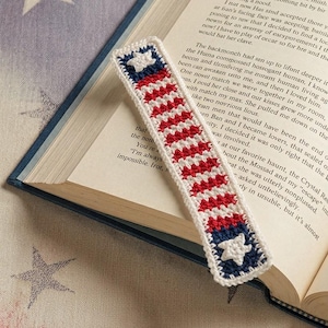 May include: A rectangular crochet bookmark featuring a pattern of red and white horizontal stripes with a dark blue section at each end containing a white star. The bookmark is crafted with a white border and is placed on an open book page.