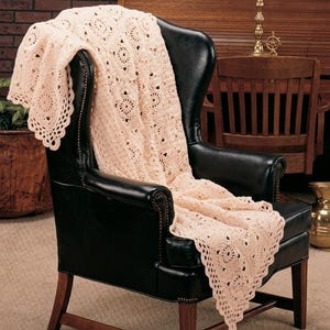 May include: A pale pink crochet blanket featuring a repeating square motif with circular centers and a scalloped edge, draped over a black leather wingback chair with brass nailhead trim and dark wooden legs.