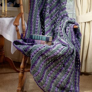 May include: A crocheted blanket in purple and grey is draped over a wooden chair. The blanket has a textured pattern with stripes of colour. Two books rest on the chair beneath the blanket. The image is well-lit, showcasing the details of the yarn.