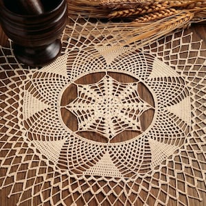 May include: A round, cream-colored crochet doily featuring an intricate, star-shaped central pattern surrounded by concentric geometric mesh designs. The delicate, hand-crafted textile displays a complex, open-work structure with pointed edges.
