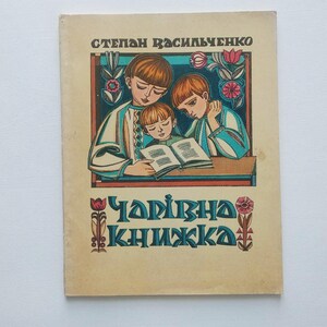 Puede incluir: Una portada de libro vintage con el título "Чарівна книжка" en escritura azul y naranja. La portada presenta una ilustración de tres niños leyendo un libro, con elementos de borde florales y decorativos. El nombre del autor, "Степан Василченко", está en la parte superior.