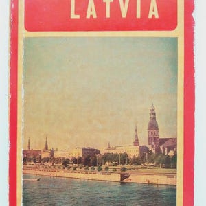 May include: Vintage travel book cover featuring the word "LATVIA" in white capital letters on a red background, with "USSR" above. The image inside shows a cityscape with a river and buildings.