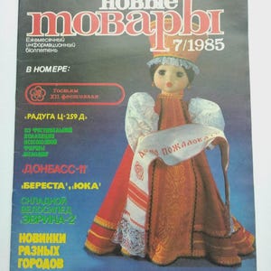 Puede incluir: Una portada de revista con una muñeca vestida con traje tradicional ruso. El título "Новые Товары" es rojo, con el número de edición 7/1985. La muñeca lleva un vestido rojo y sostiene una pancarta con texto.