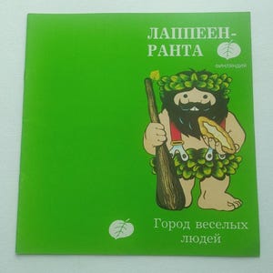 Puede incluir: Postal verde con un personaje de dibujos animados barbudo, sosteniendo un bastón y un pastel. El personaje tiene una corona de hojas y ropa. El texto ruso incluye "Lapenranta" y "Ciudad de la gente feliz".