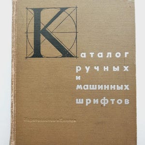 May include: A vintage book with a tan cover featuring a large black letter "K" inside a circle with intersecting lines. The title is in white Cyrillic text. The book appears to be a catalog of fonts.