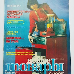 Puede incluir: Portada de revista con una mujer con camisa roja y delantal, mostrando un electrodoméstico de cocina y una batidora. El título de la revista es "Nuevos productos" con el número de edición 6/1985. También se ve texto en ruso.