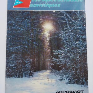 Op de afbeelding: Een vintage reisfoldercover met een besneeuwd boslandschap met zonlicht dat door de bomen schijnt. Het bovenste gedeelte heeft tekst in turquoise en rood, met het jaartal "1982". De onderkant heeft het Aeroflot-logo en "Soviet airlines".
