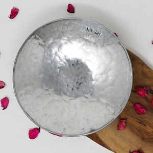 May include: A silver hammered metal bowl with a textured surface. The bowl is sitting on a wooden cutting board with pink flower petals scattered around it. The bowl has the text "HHH" inscribed on the bottom.