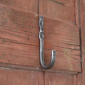 Puede incluir: Un gancho de metal forjado a mano montado sobre una superficie de madera rojiza y envejecida. El gancho es de color gris oscuro y tiene un diseño curvado. La madera tiene vetas y nudos visibles, lo que añade a su aspecto rústico.
