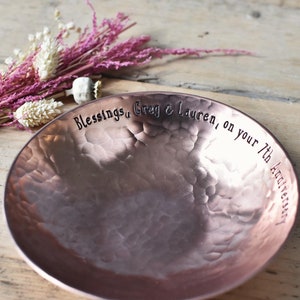 A bright hammered copper bowl with an inscription on the inside rim that reads, 'Blessings, Greg & Lauren, on your 7th Anniversary'. The hand crafted bowl sits on a wooden slatted table next to a small pink posy of dried flowers.
