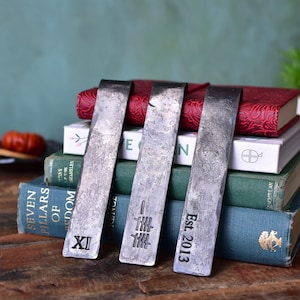 May include: Three silver metal bookmarks with different engraved designs. The bookmarks are placed on top of a stack of books. The designs include Roman numerals, a series of lines, and the text "Est. 2013".