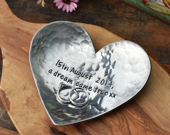 May include: A silver heart-shaped dish with a hammered texture. The dish has the inscription "15th August 2014, a dream came true xx" and two rings are resting inside.