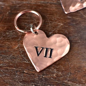 Op de afbeelding: Een koperen sleutelhanger in de vorm van een hart met een gehammerde textuur en het Romeinse cijfer VII gegraveerd in zwart.